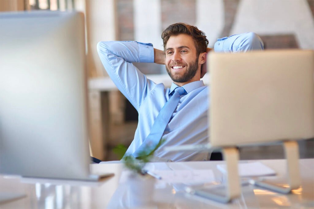 “Man sitting at desk with relaxed posture, smiling, hands behind head, enjoying stress-free work environment with computer screens on table.”