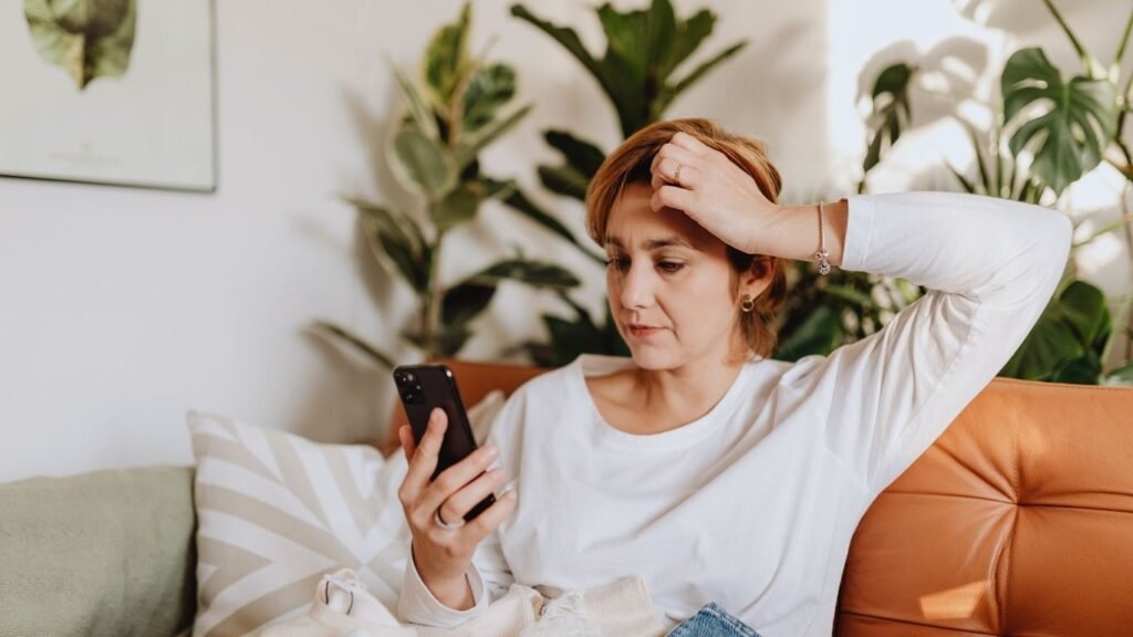 A midlife woman sitting on a sofa, looking at her phone with a thoughtful expression, resting her hand on her head in a calm home setting.