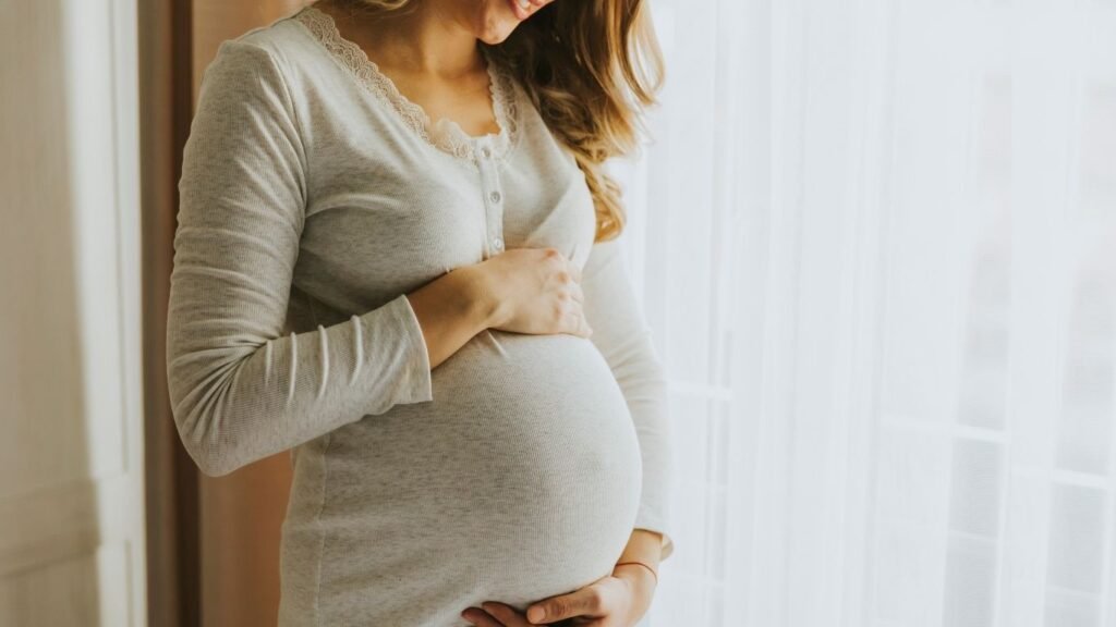 A pregnant woman gently holding her belly while standing near a window in soft natural light.
