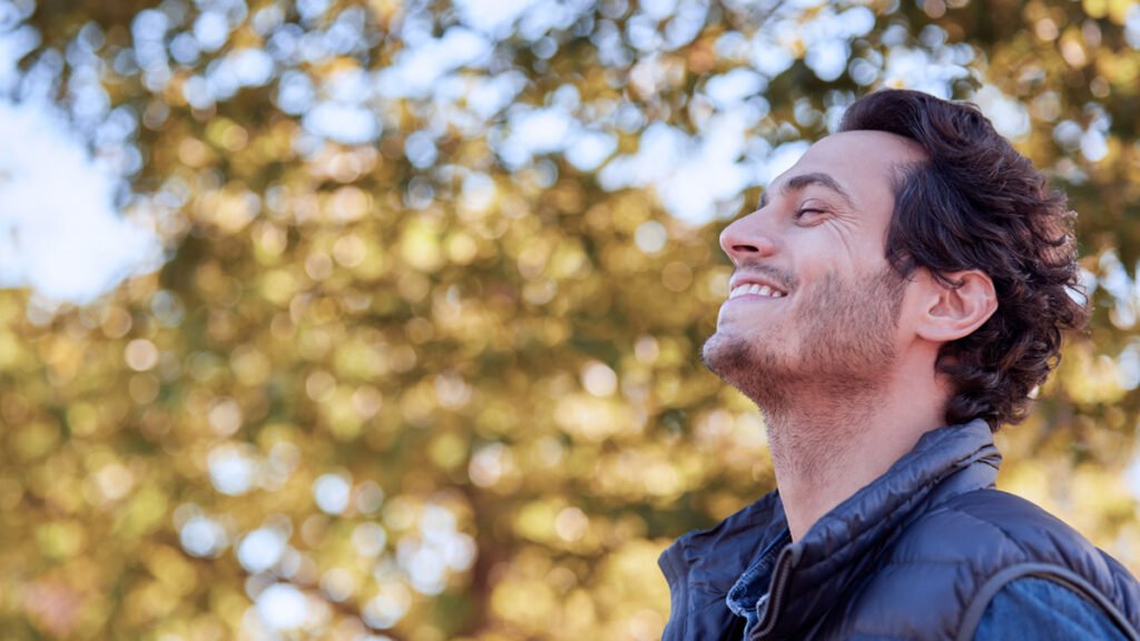A man standing outdoors with eyes closed, breathing deeply, surrounded by trees in soft natural light.